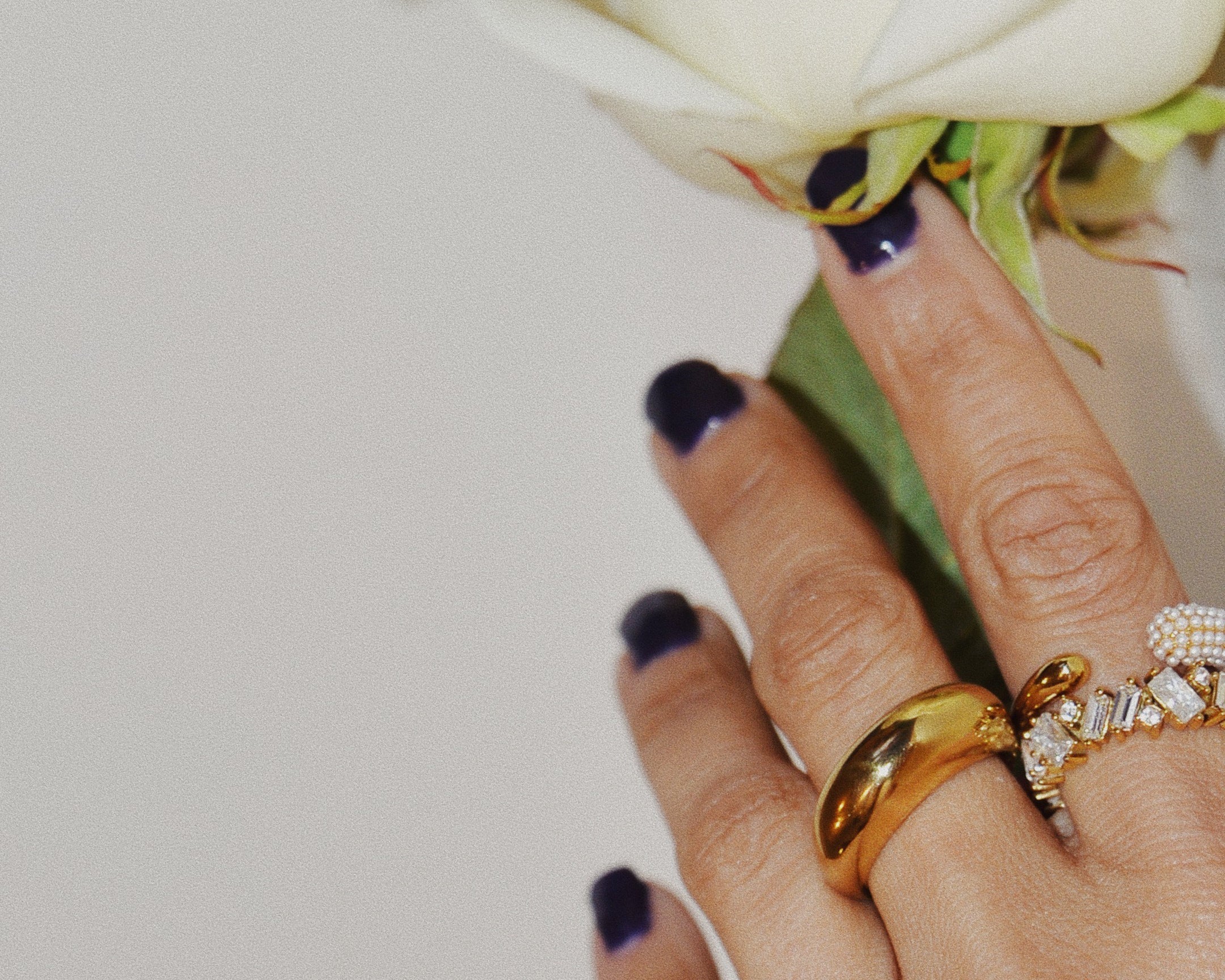 Hands holding a white flower with dark blue nails and the Lisa Gold Crystal Gold Ring with another two rings